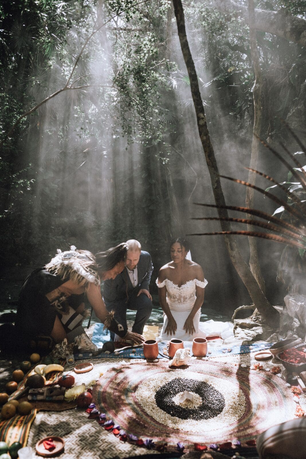 Couple kneels at the sacred jungle altar, asking the land and the ancestors for permission to bless their union with a Mayan Shaman at their side. Beams of light and smoke cut through the jungle canopy at this Mayan Elopement Ceremony at a cenote in Tulum.