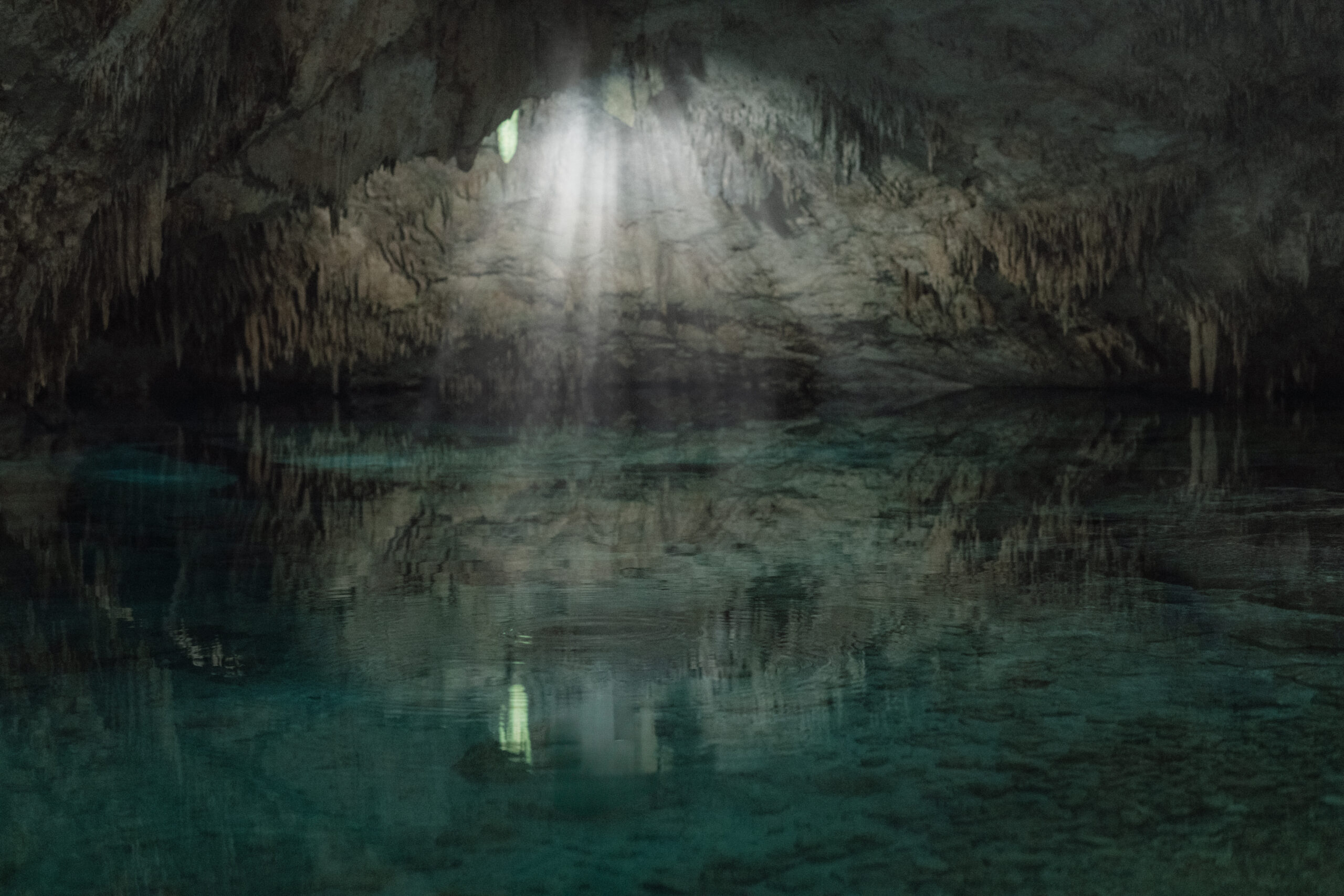 Cenote in Tulum, Mexico with sunlight streaming through an opening into crystal clear water, creating light beams inside a cave, representing a sacred and spiritual elopement location.